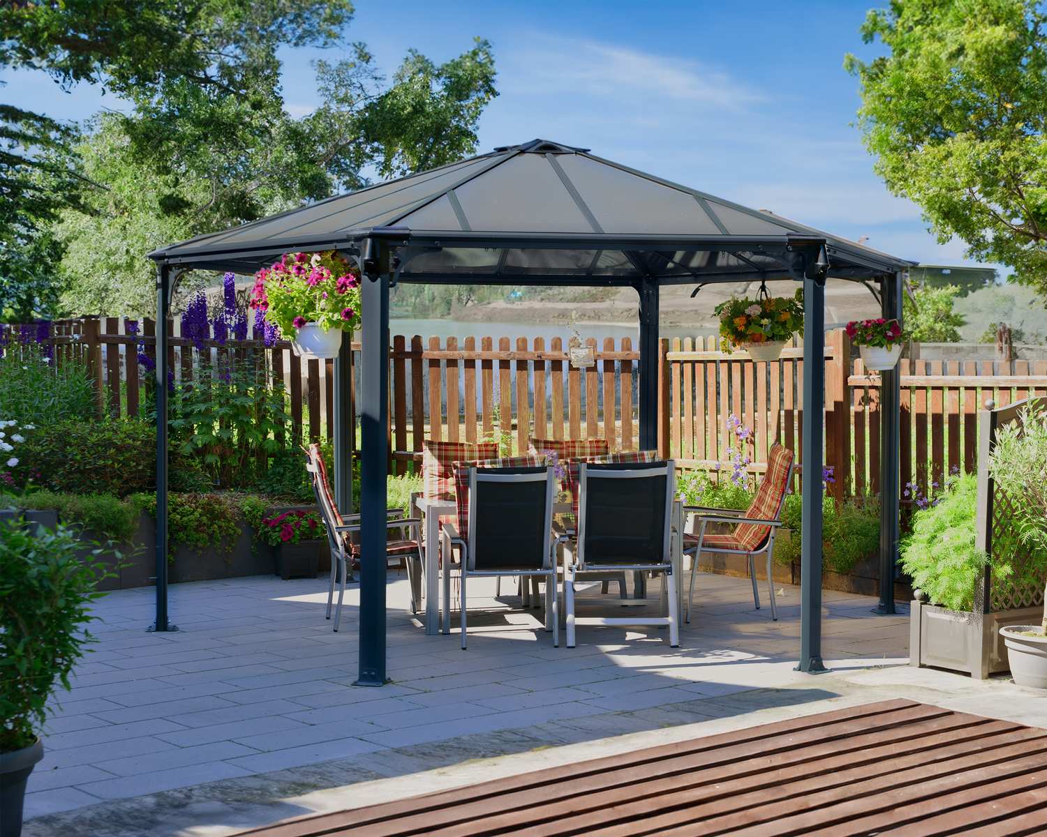 Monaco 13ft x 15ft grey hexagonal aluminum gazebo with wooden table and chairs on a paved patio, set in a lush garden at sunset