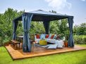 Palermo 12ft x 12ft grey aluminum gazebo with curtains, a sofa with colorful cushions, and a wooden deck in a green garden