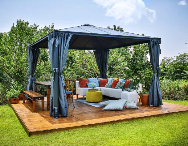 Palermo 12ft x 12ft grey aluminum gazebo with curtains, a sofa with colorful cushions, and a wooden deck in a green garden