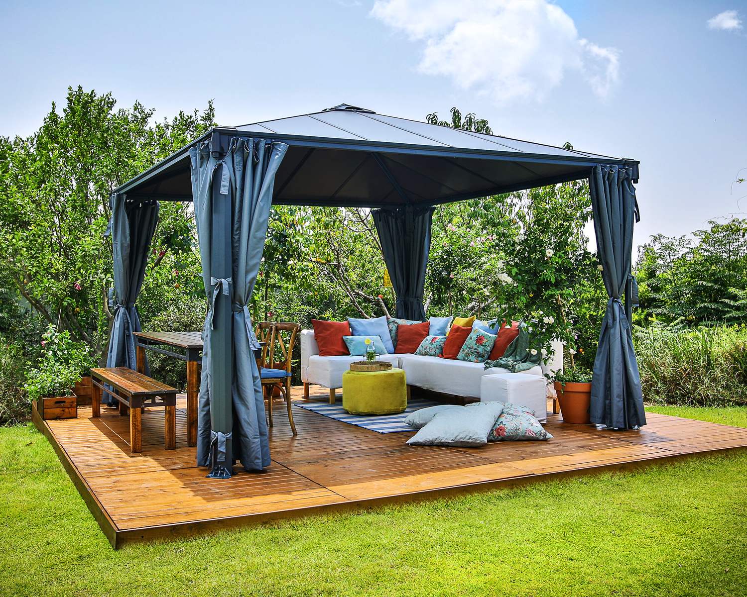 Palermo 12ft x 12ft grey aluminum gazebo with curtains, a sofa with colorful cushions, and a wooden deck in a green garden