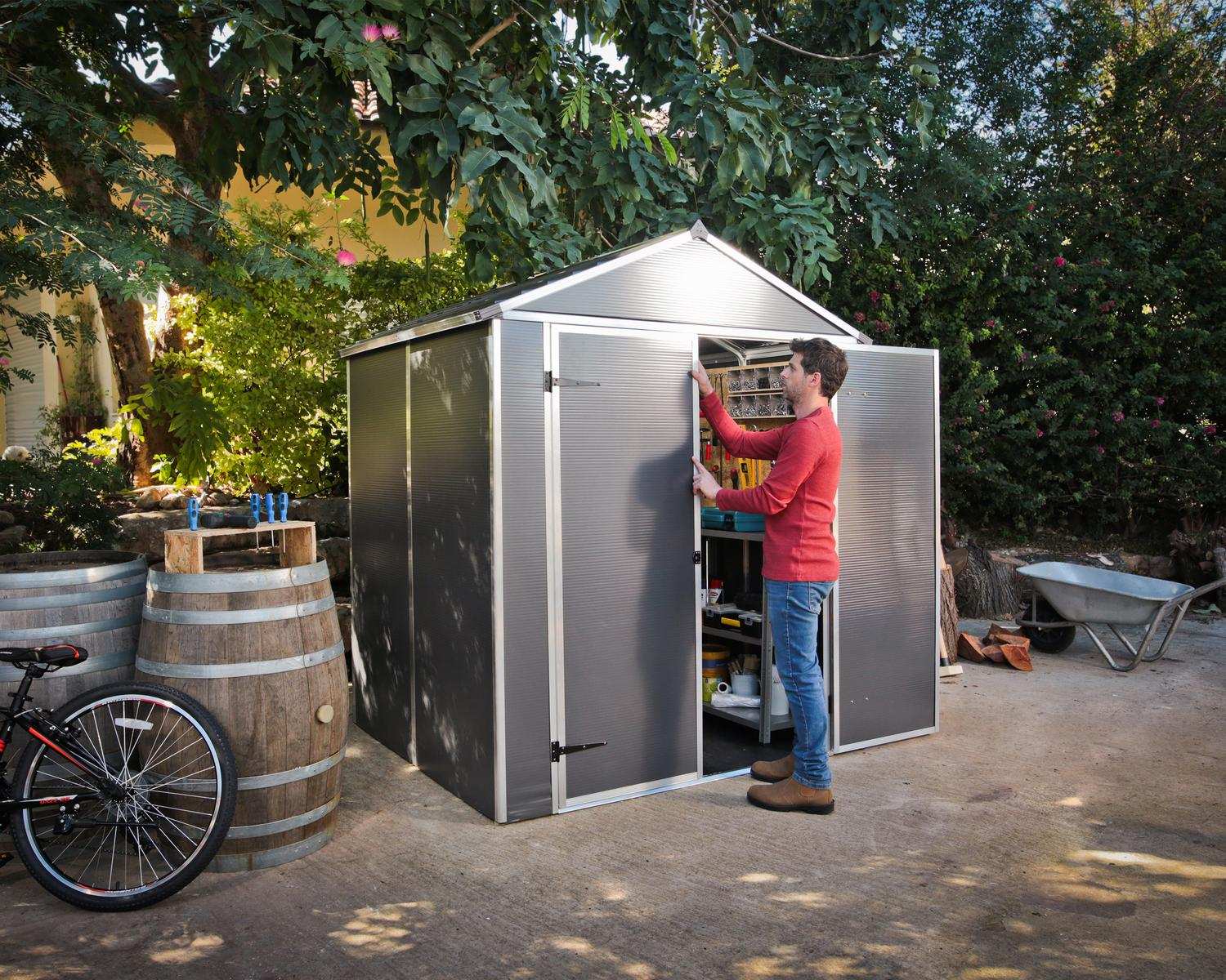 Dark grey Rubicon plastic storage shed, 6x5 ft, with double doors and a sloped roof, on grass surrounded by plants