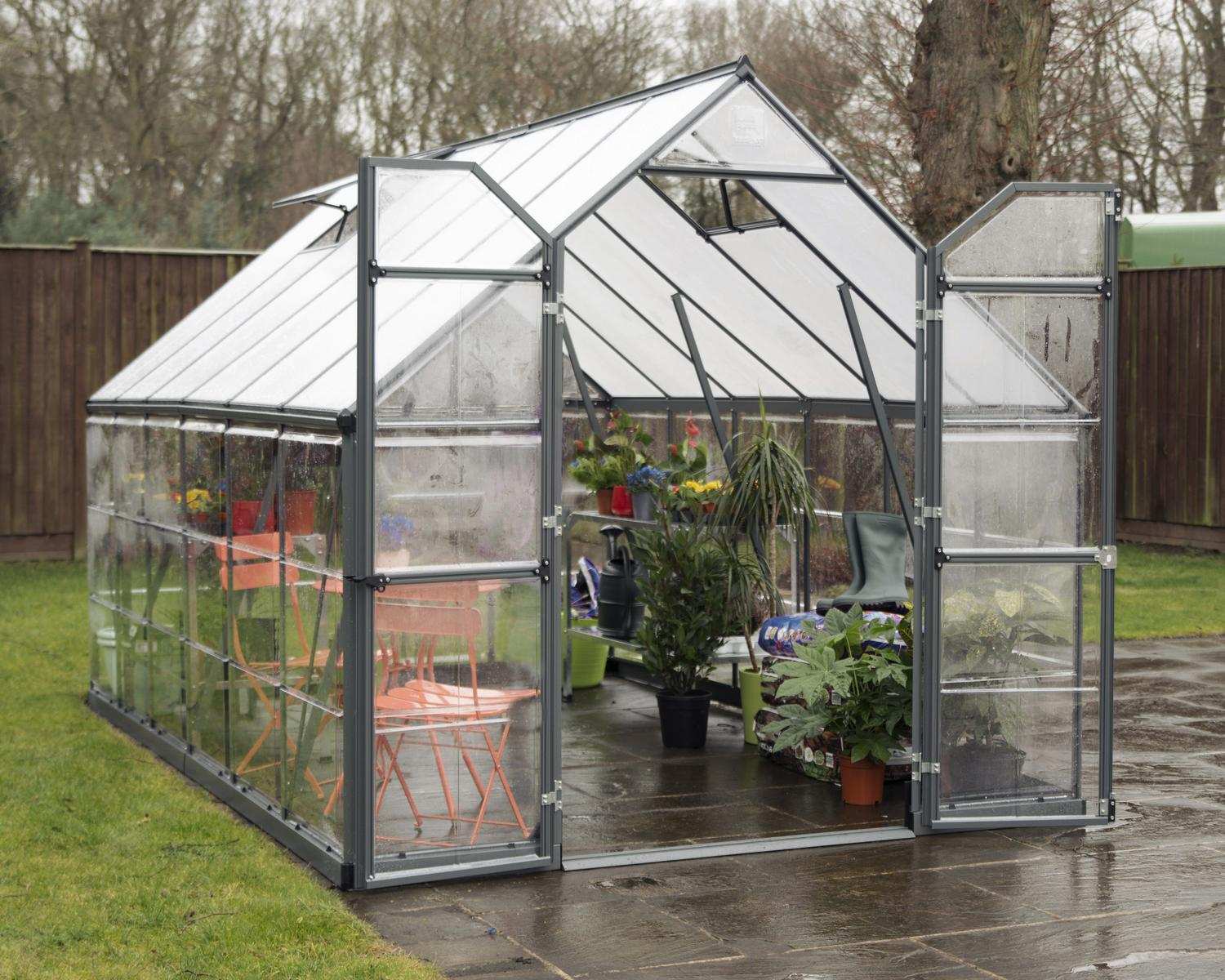 Balance 8ft x 12ft grey greenhouse with clear polycarbonate panels and aluminium frame, on a wet patio, surrounded by a grassy yard and fence