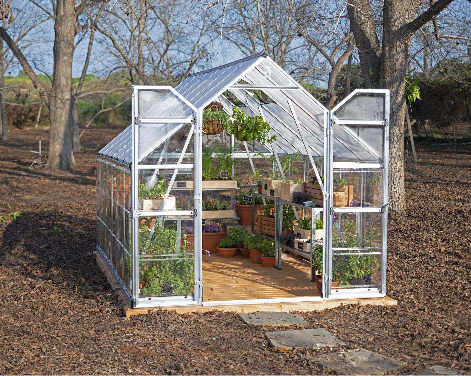 8ft x 12ft silver Balance greenhouse with polycarbonate panels and aluminium frame, surrounded by plants and a wheelbarrow