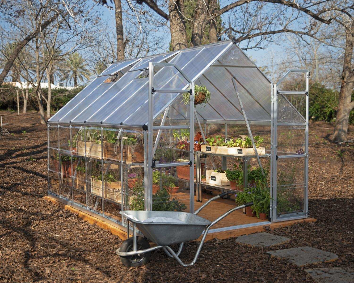 8ft x 12ft silver Balance greenhouse with polycarbonate panels and aluminium frame, surrounded by plants and a wheelbarrow