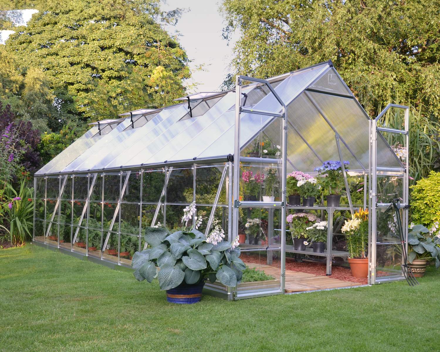 Balance 8ft x 20ft silver greenhouse with clear polycarbonate panels and aluminium frame, set in a garden with plants surrounding it
