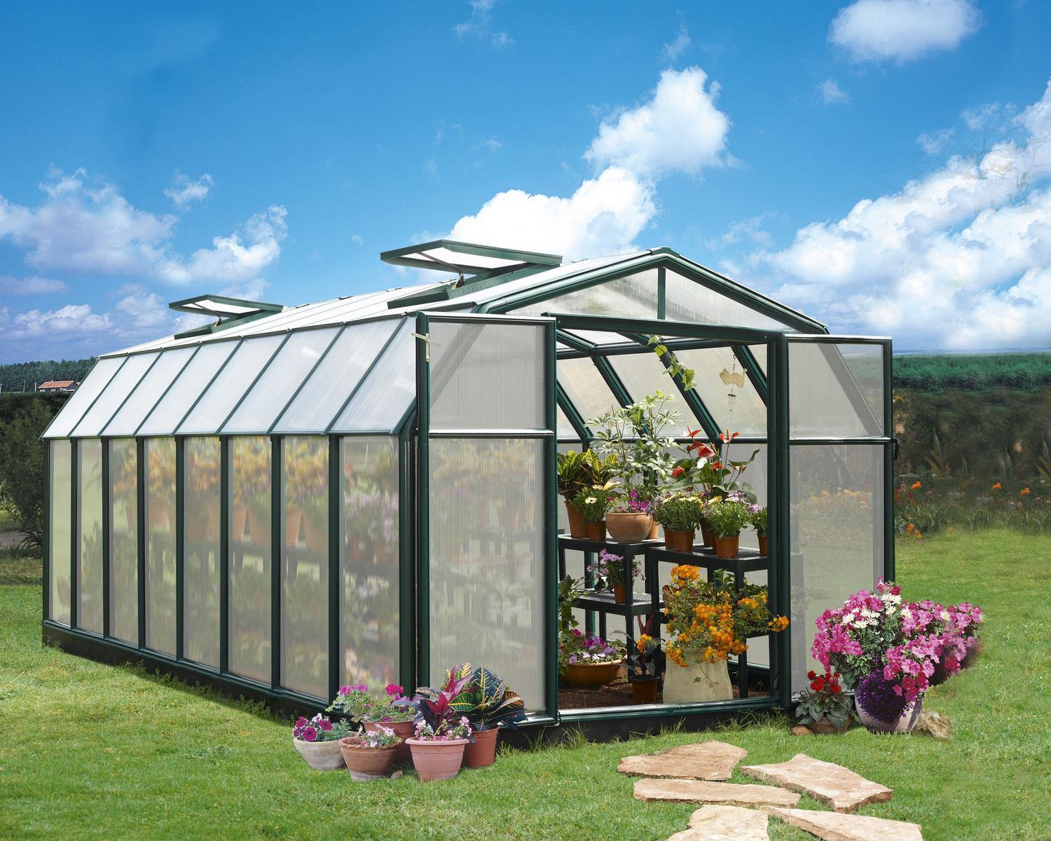 Hobby Gardener 8ft x 16ft green greenhouse with polycarbonate panels, brimming with plants inside and out, beneath a blue sky