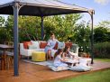 Palermo 12ft x 12ft grey aluminum gazebo with curtains, a sofa with colorful cushions, and a wooden deck in a green garden