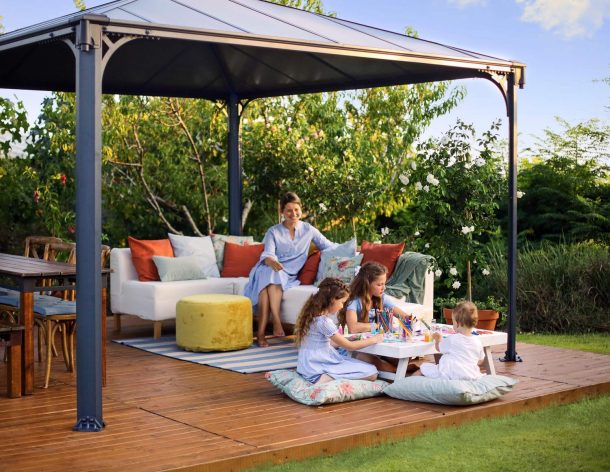 Palermo 12ft x 12ft grey aluminum gazebo with curtains, a sofa with colorful cushions, and a wooden deck in a green garden