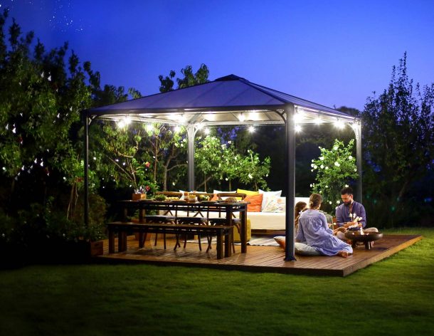 Palermo 12ft x 12ft grey aluminum gazebo with curtains, a sofa with colorful cushions, and a wooden deck in a green garden
