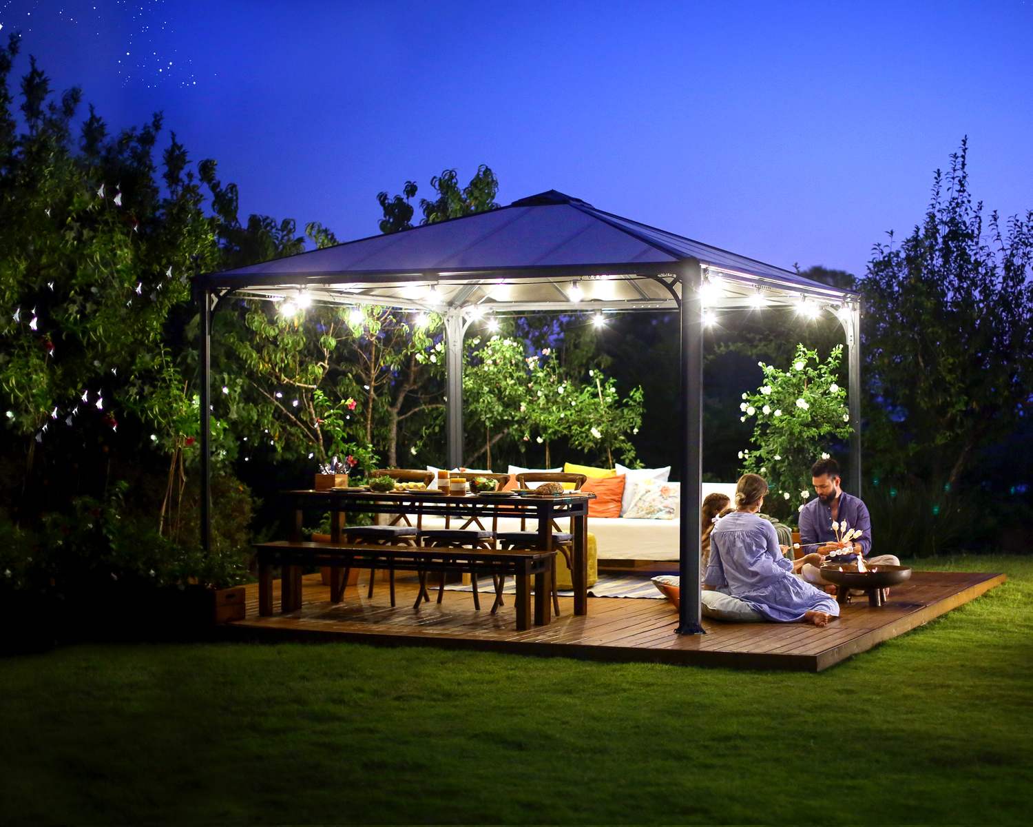 Palermo 12ft x 12ft grey aluminum gazebo with curtains, a sofa with colorful cushions, and a wooden deck in a green garden