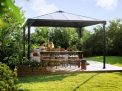 Palermo 12ft x 12ft grey aluminum gazebo with curtains, a sofa with colorful cushions, and a wooden deck in a green garden