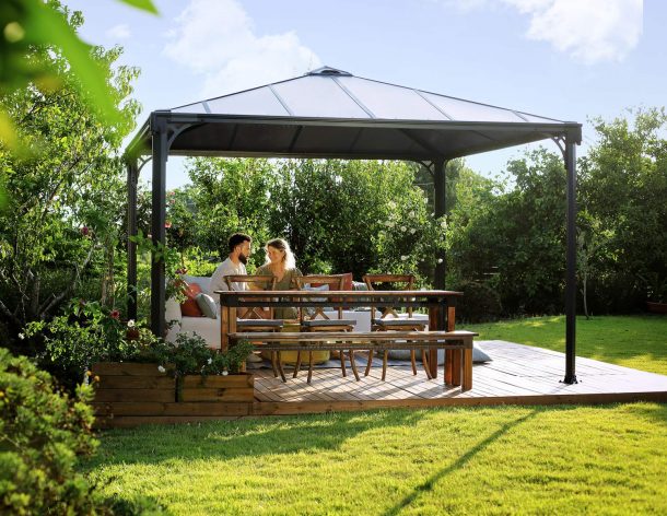 Palermo 12ft x 12ft grey aluminum gazebo with curtains, a sofa with colorful cushions, and a wooden deck in a green garden