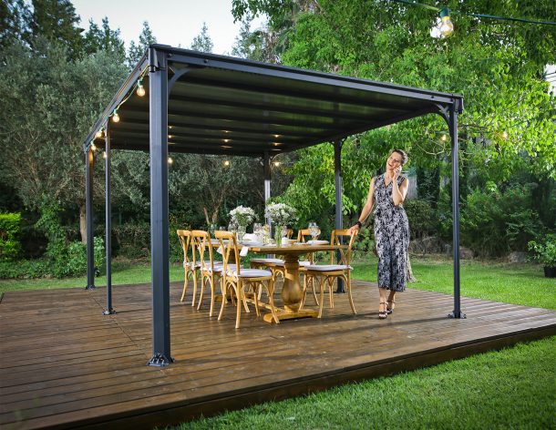 A flat roof gazebo 10&#039; x 14&#039; covers a deck patio with dining furniture and a lady talking on the phone