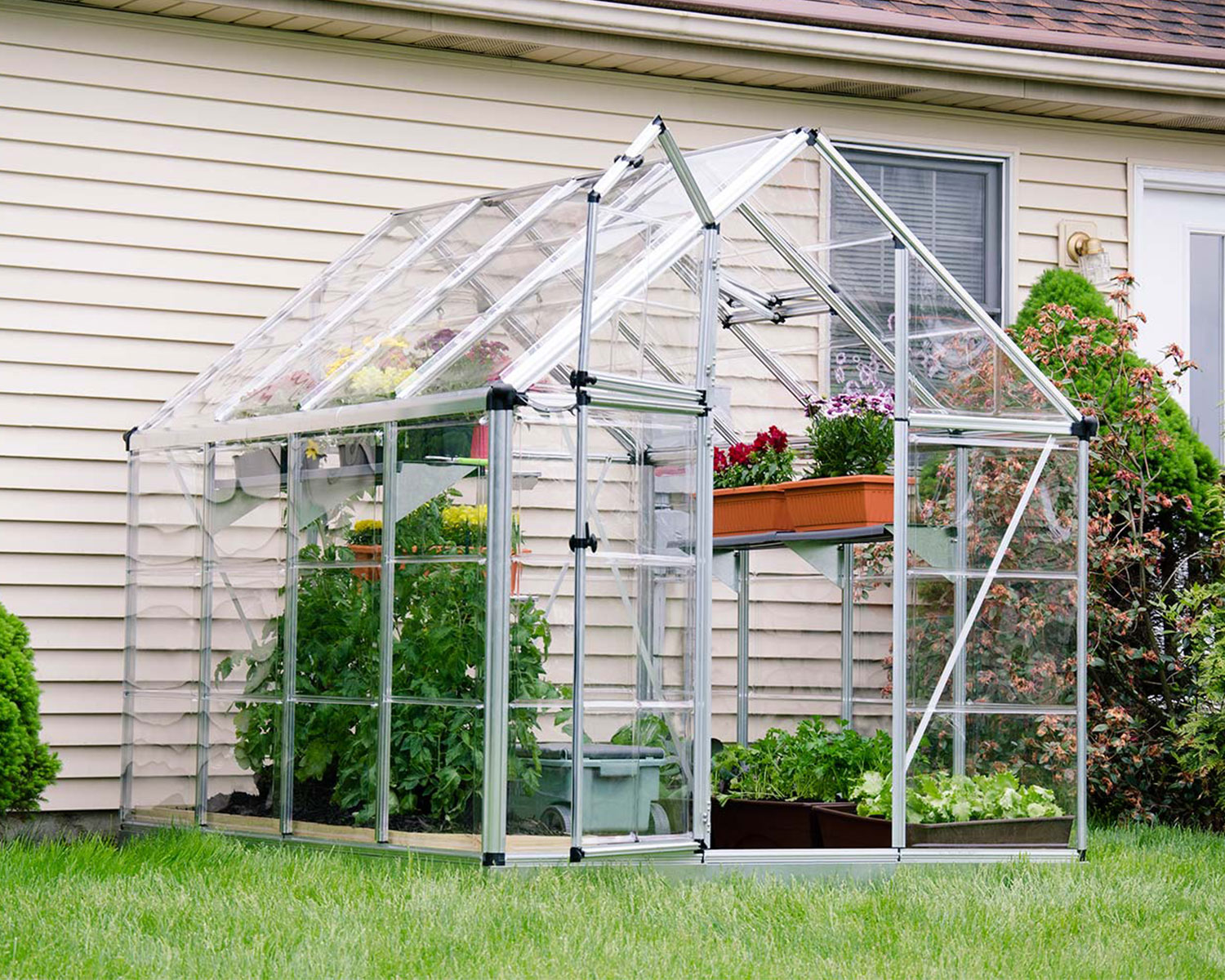 Snap &amp; Grow 6 ft. x 8 ft. Greenhouse Silver Structure &amp; Clear Panels on a lawn next to a house