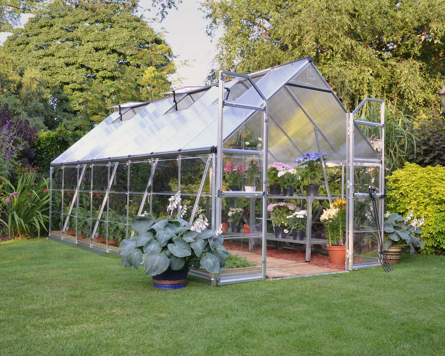 8ft x 16ft silver Balance greenhouse with polycarbonate panels and a sturdy aluminium frame, filled with plants, set in a lush garden with a green lawn
