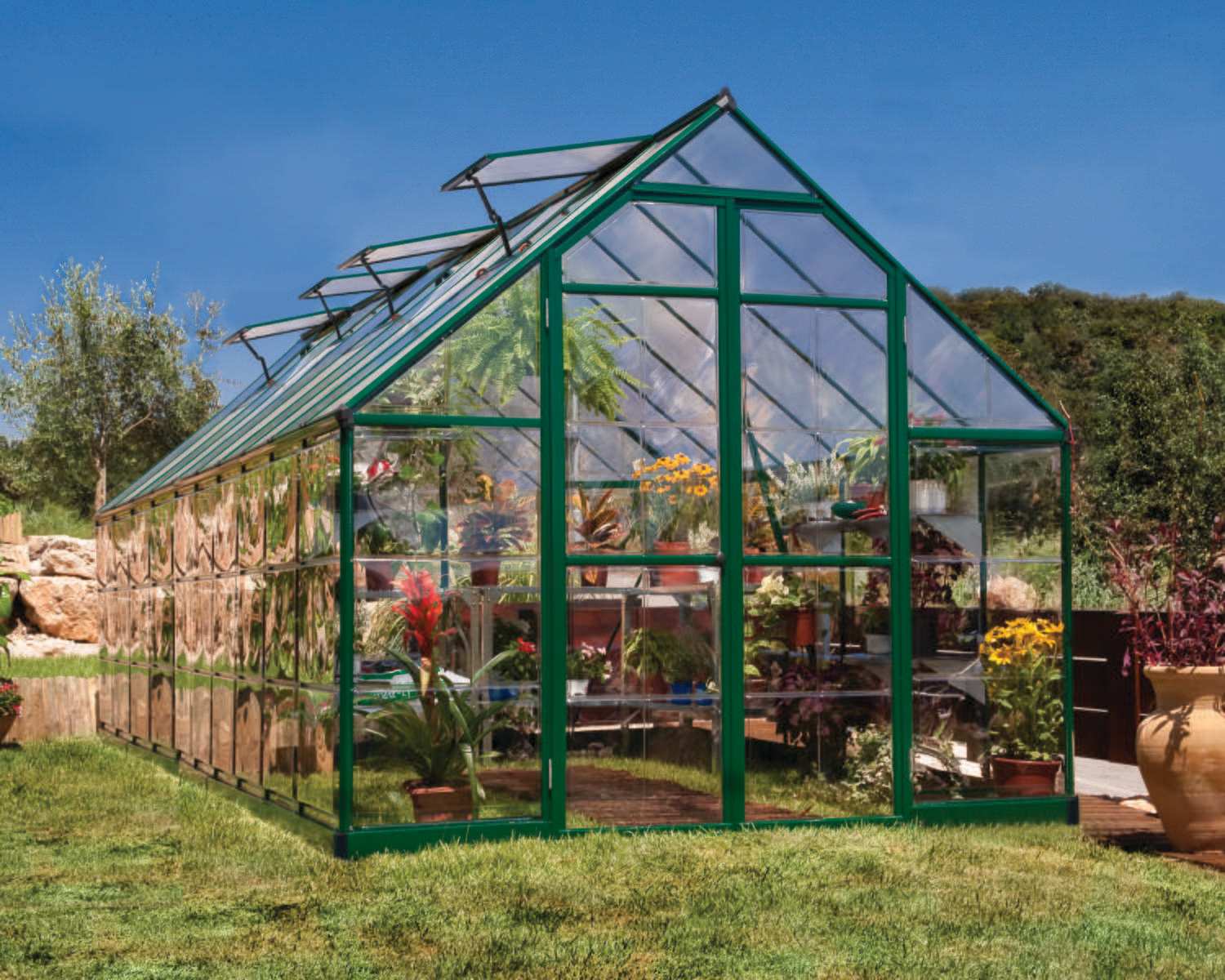 Balance 8ft x 20ft green greenhouse with clear polycarbonate panels and aluminium frame, set in a sunlit garden full of plants