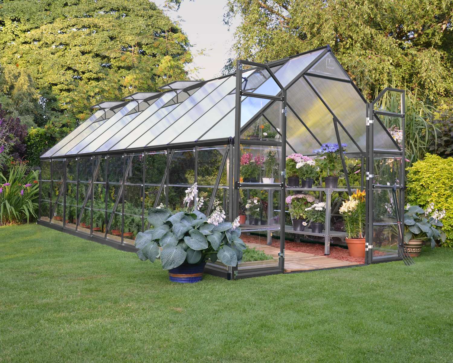 Balance 8ft x 20ft grey greenhouse with clear polycarbonate panels and aluminium frame, set in a vibrant garden with lush plants