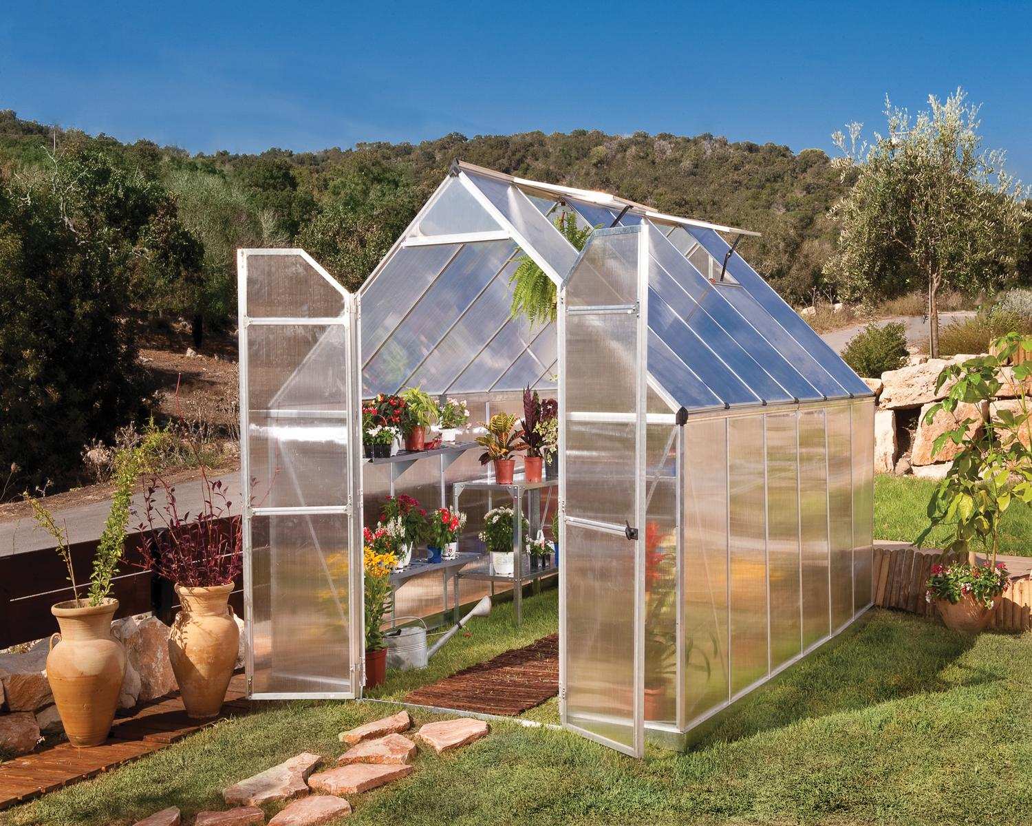 Essence 8x12 Silver greenhouse with polycarbonate panels, double doors, and a sloped roof, set in a vibrant garden