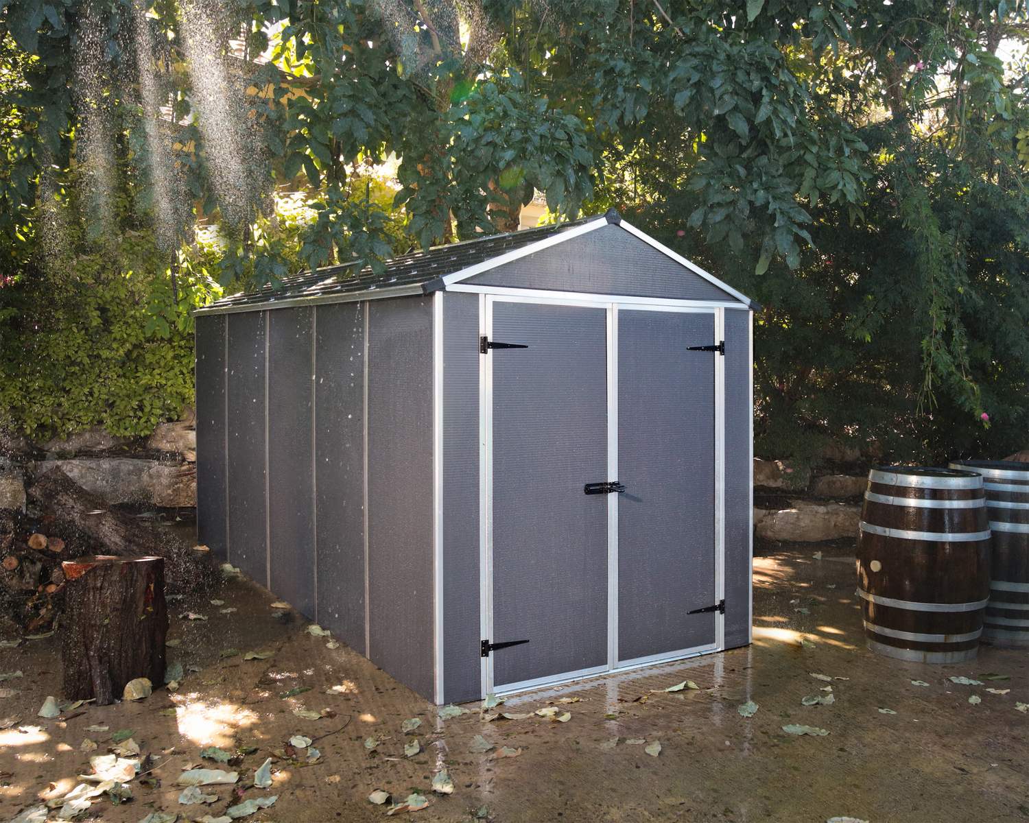 6ft x 12ft grey Rubicon garden shed with a sturdy aluminum frame and multiwall polycarbonate panels, set among plants with a wooden barrel