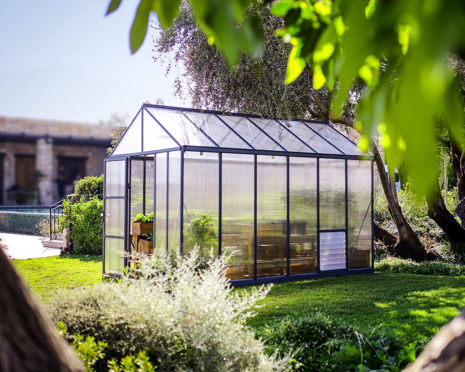 8ft x 12ft grey Glory greenhouse with a durable aluminum frame and multi-wall polycarbonate panels, brimming with plants, nestled in a lush garden