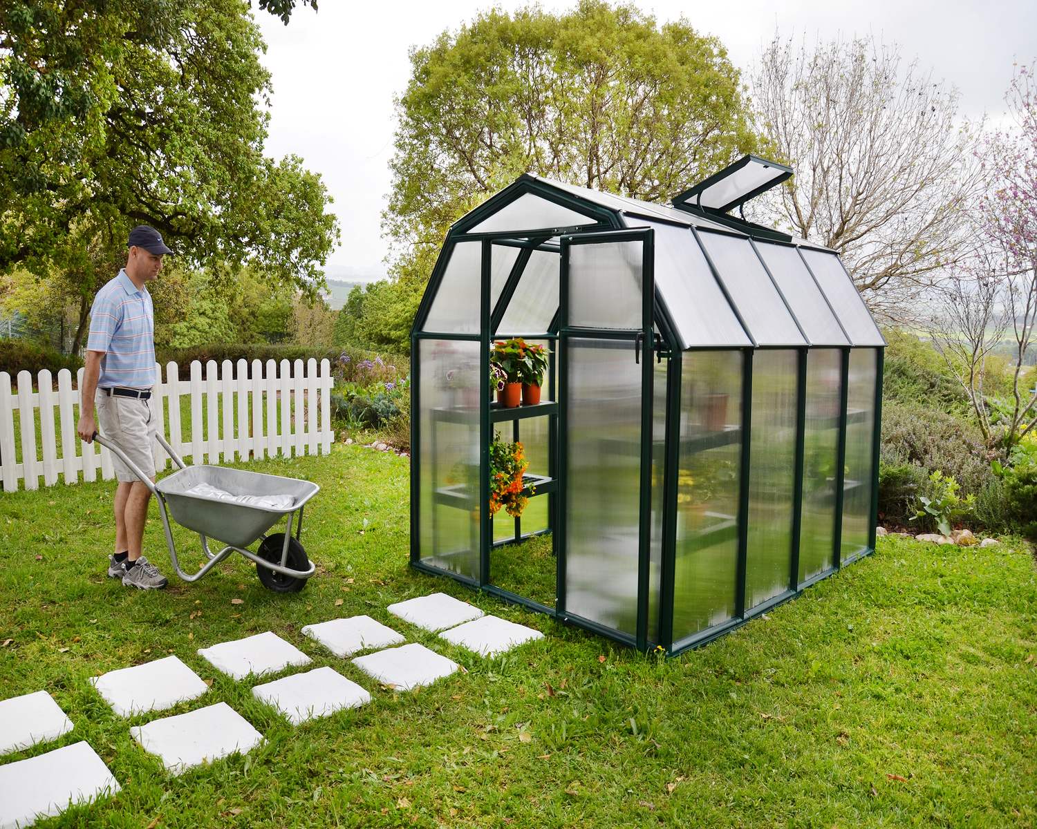 6ft x 8ft green EcoGrow greenhouse with a durable aluminum frame and clear polycarbonate panels, set in a vibrant garden with plants around it