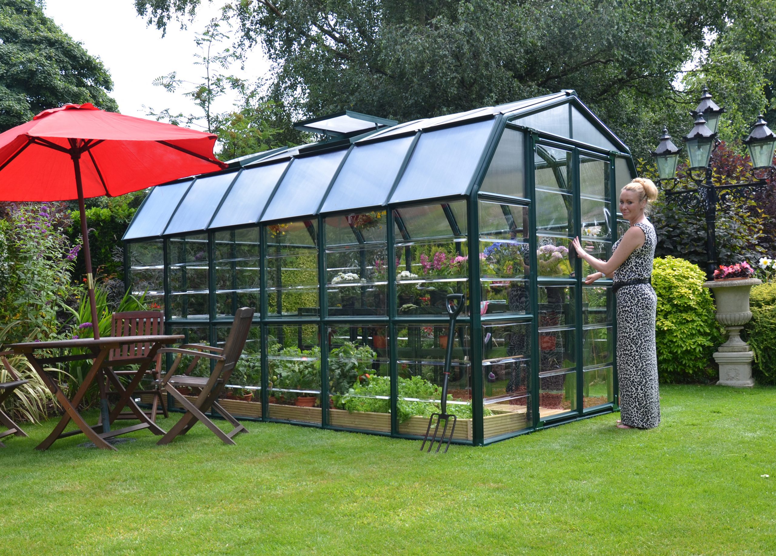 8ft x 12ft green Grand Gardener greenhouse with clear polycarbonate panels, in a garden with a red umbrella and wooden chairs