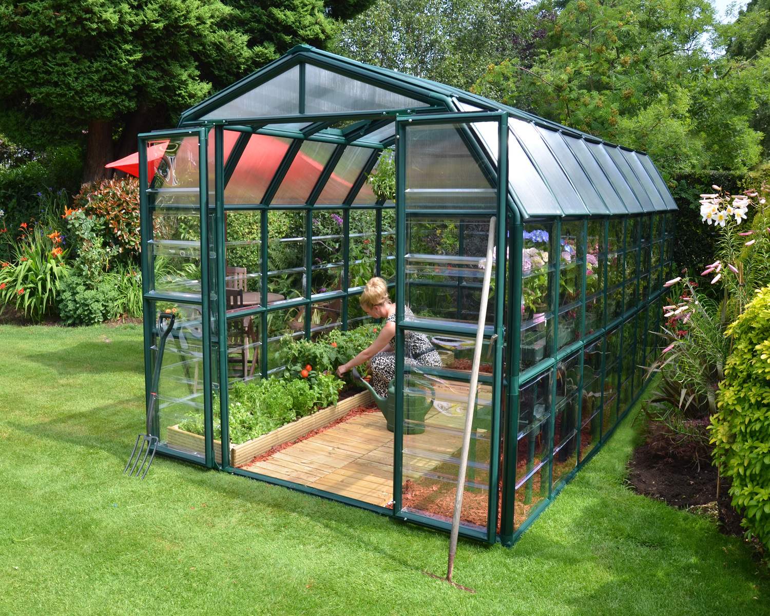 8ft x 16ft green Grand Gardener greenhouse with clear polycarbonate panels, surrounded by plants, a wooden table, and a red umbrella in a lush garden