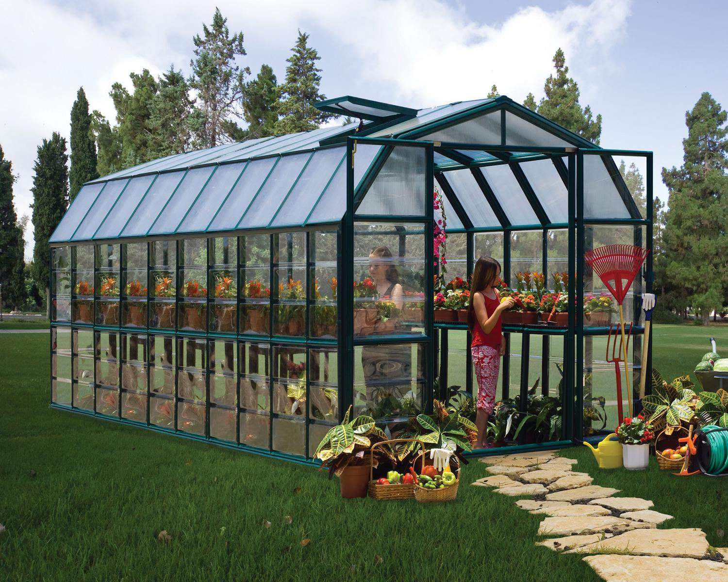 8ft x 20ft green Grand Gardener greenhouse with clear polycarbonate panels and an aluminum frame, with plants in a lush garden
