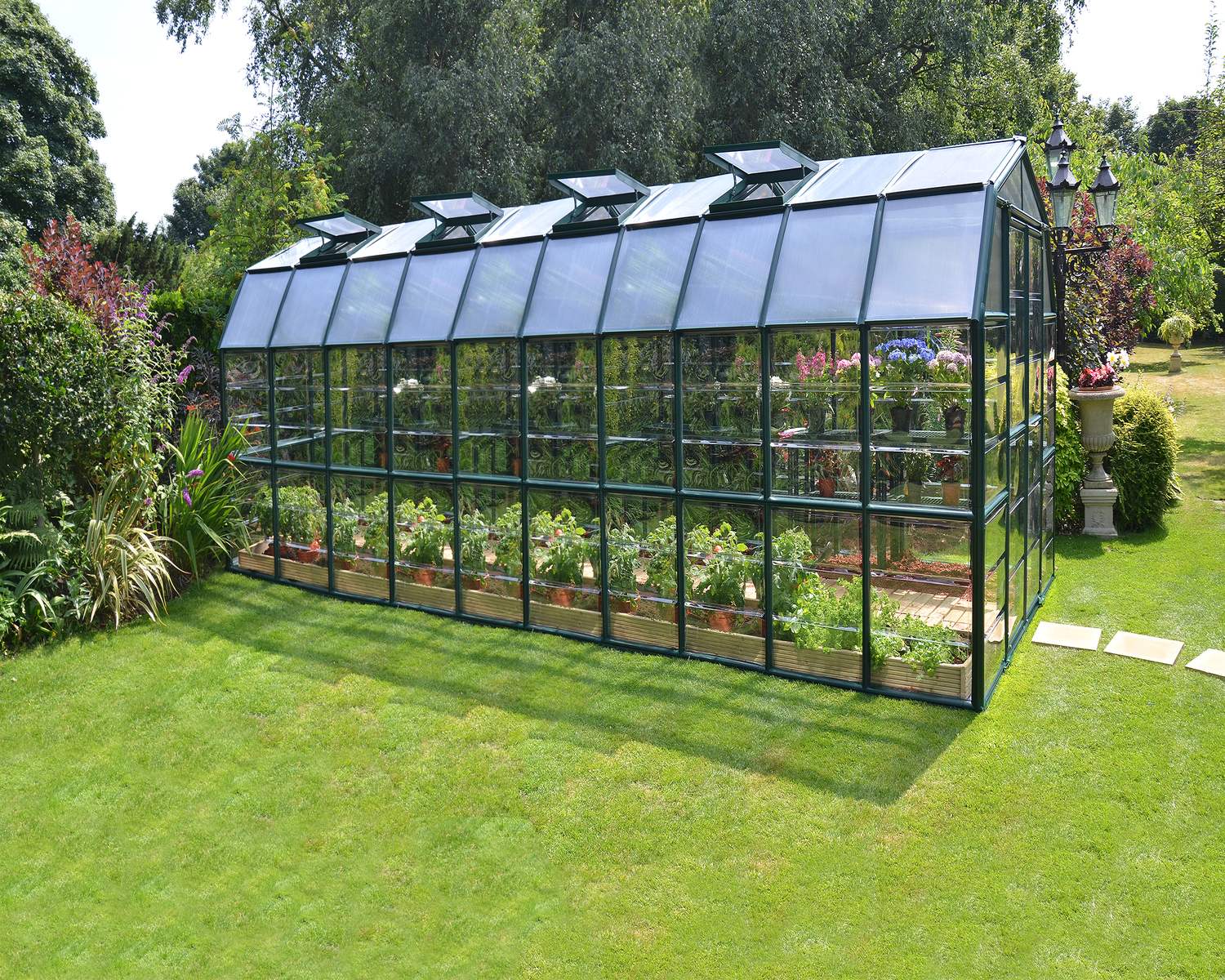 8ft x 20ft green Grand Gardener greenhouse with clear polycarbonate panels and an aluminum frame, with plants in a lush garden