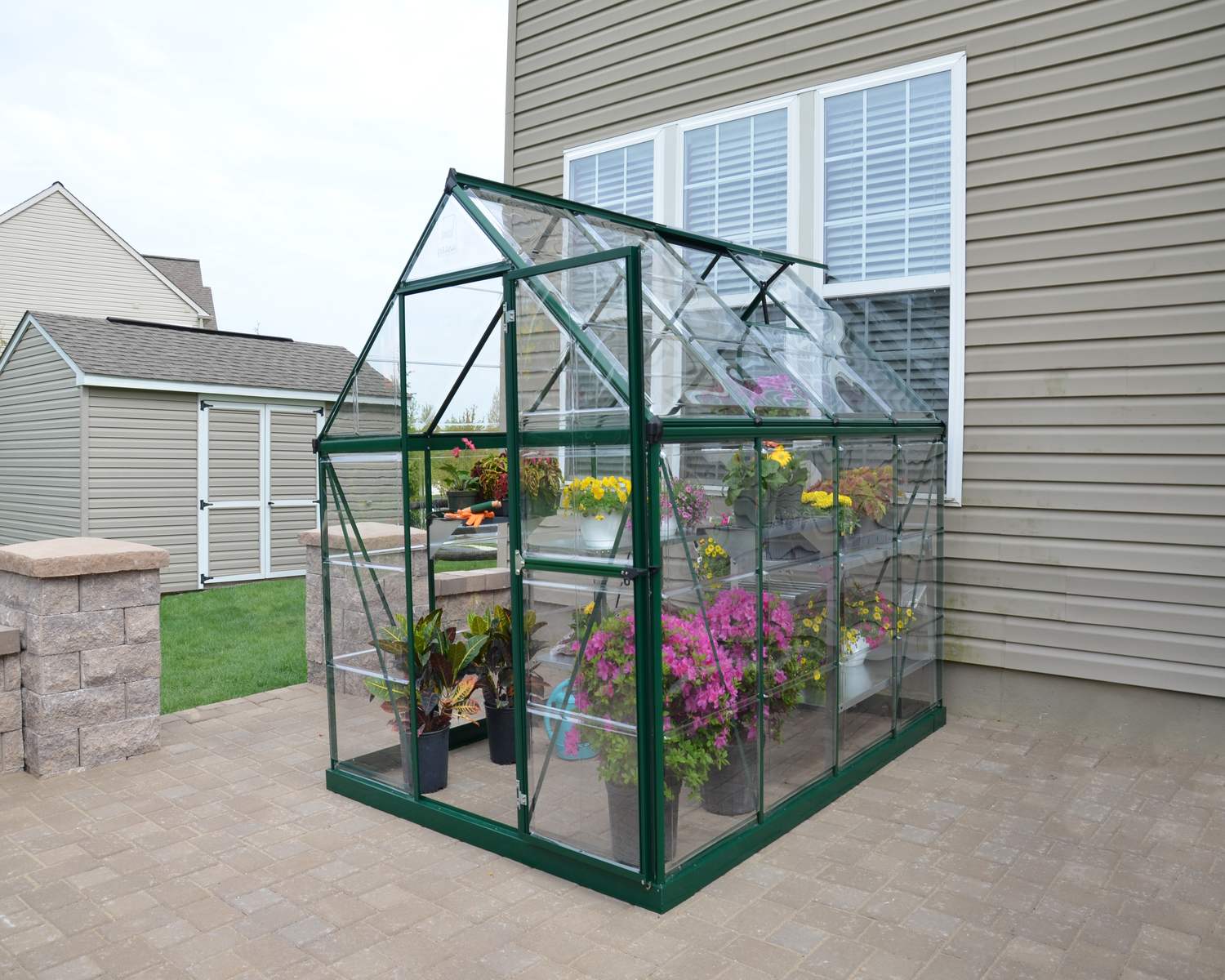 6ft x 8ft green Harmony greenhouse with clear polycarbonate panels and an aluminum frame, set on a residential patio near a beige house