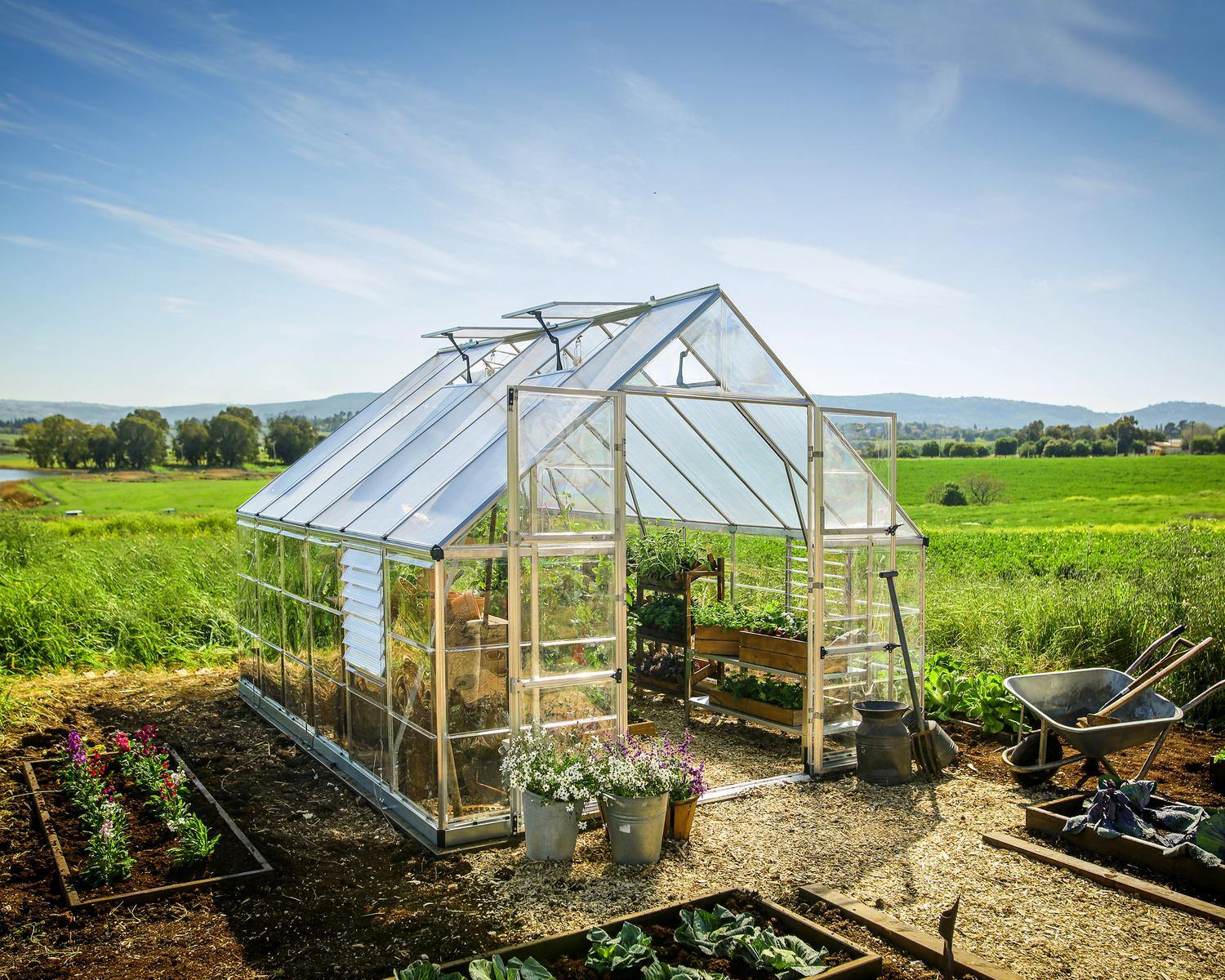 10ft x 12ft silver Balance greenhouse with aluminum frame and clear polycarbonate panels, brimming with plants and surrounded by lush greenery