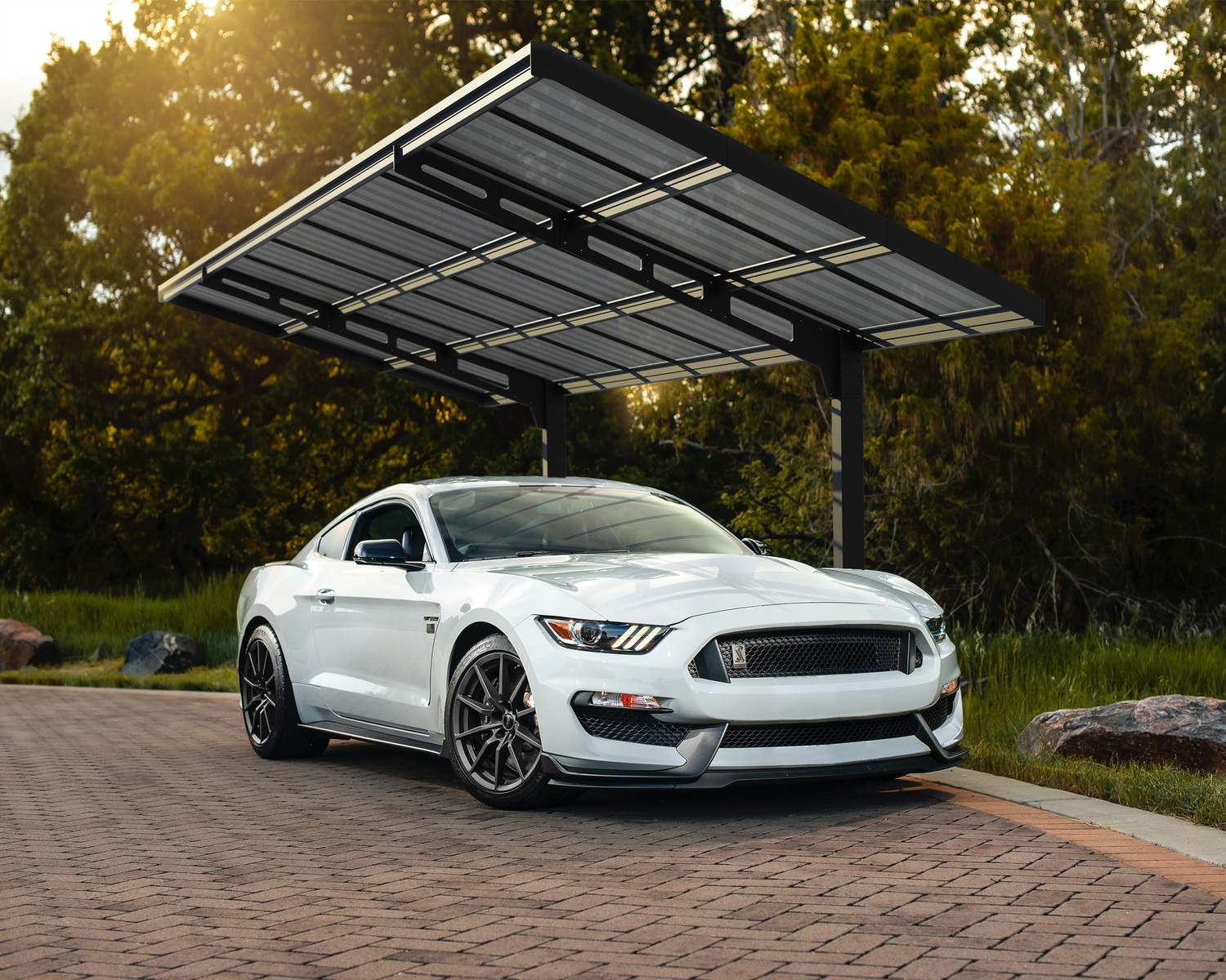 10ft x 20ft Sydney Wave carport with a black aluminum frame and polycarbonate roof, sheltering a white sports car, surrounded by greenery