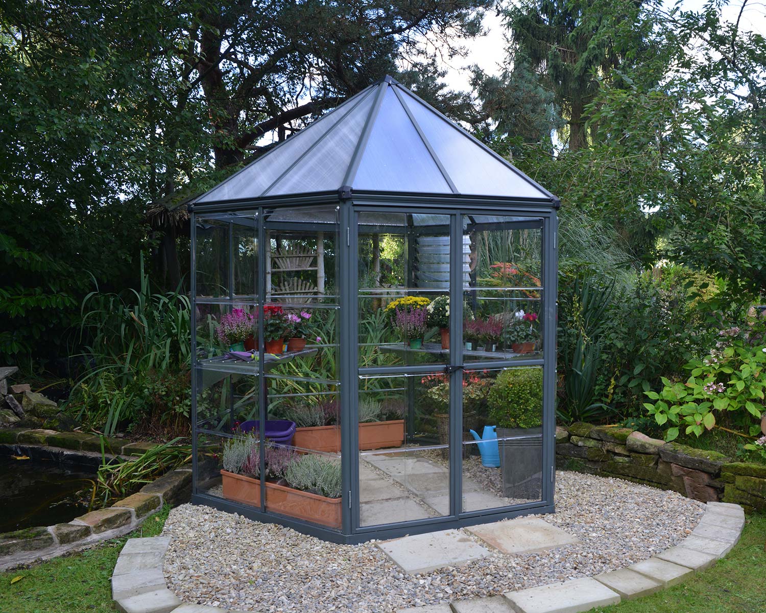 8ft grey Oasis greenhouse with an aluminum frame and clear polycarbonate panels, placed in a vibrant garden setting with lush greenery